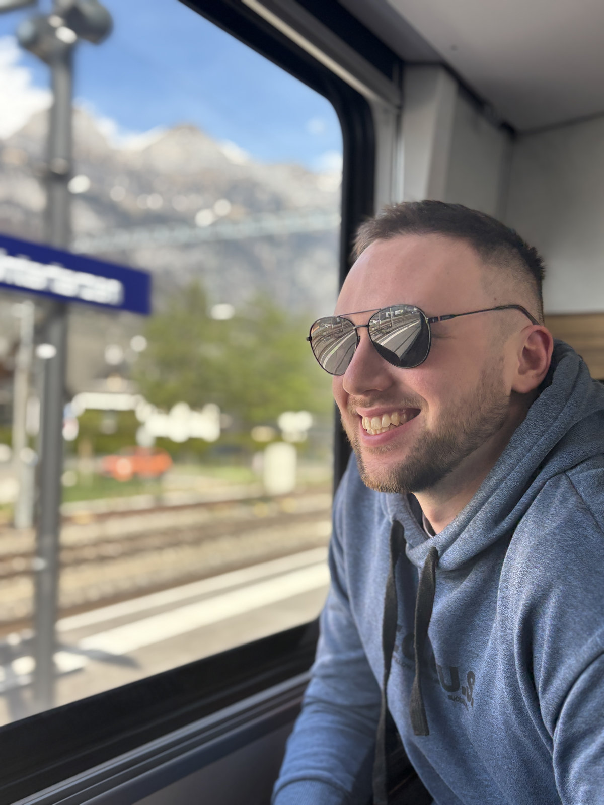 Railhop founder smiling out a train window with Swiss mountains and Interlaken station in the background